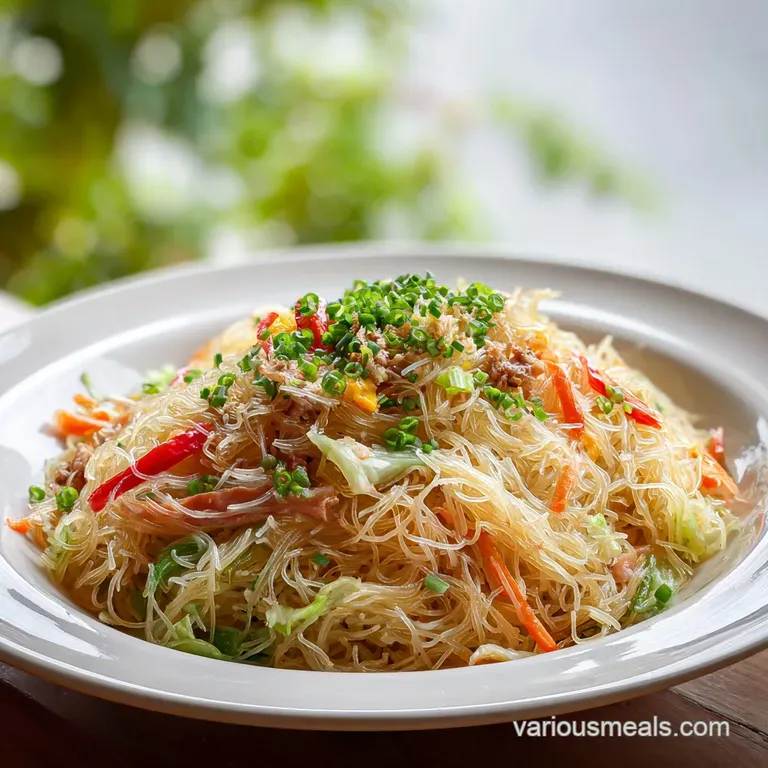 Pancit piled high on a white plate. Steaming noodles catch the light, dotted with colorful veggies and juicy meat.
