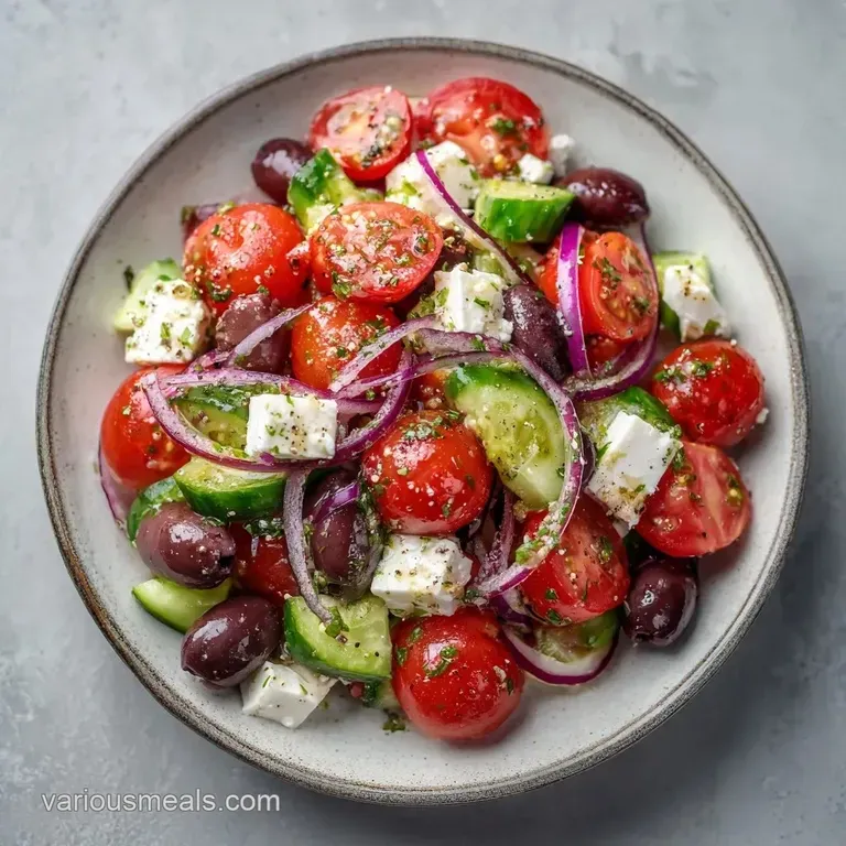 Vibrant red tomatoes and sliced cucumbers topped with creamy feta chunks and olives on a white ceramic platter.